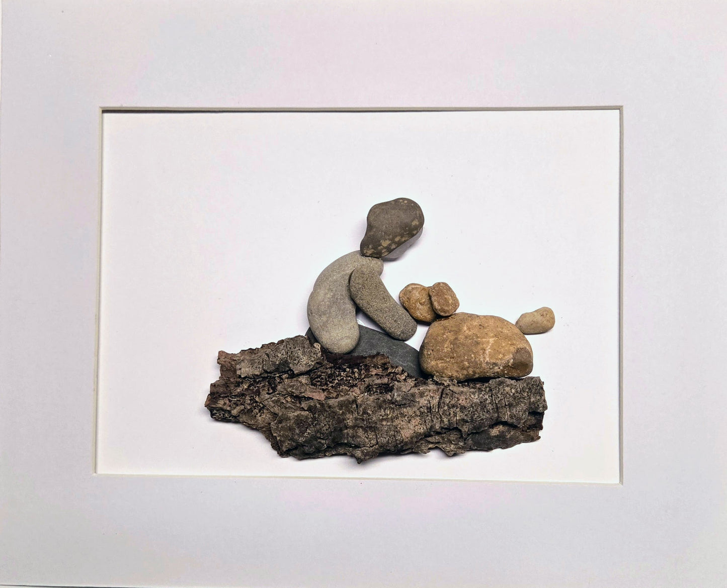 Artistic arrangement of stones shaped like a person and a dog on a log, framed in white.
