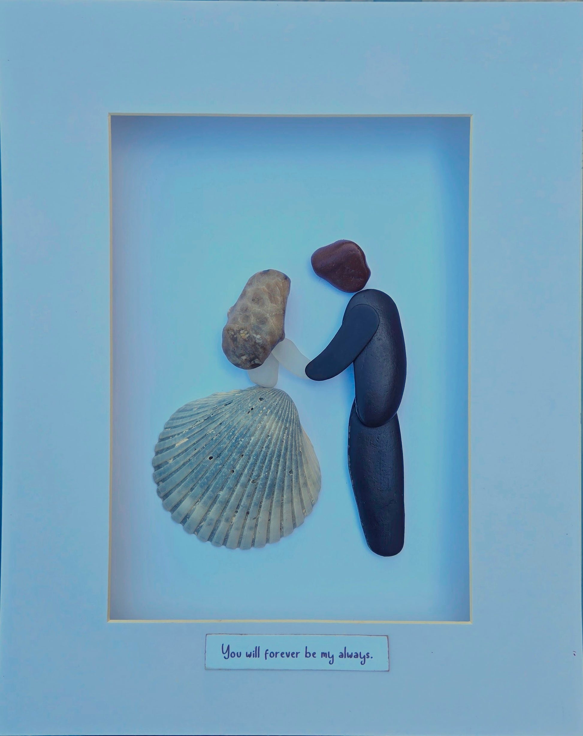 Forever My Always
A tender moment captured in stone and shell—two figures holding hands in quiet devotion. Her flowing skirt is a shell gathered from Argentina, her graceful form shaped from sea glass, and her hair a natural Petoskey stone, rich with ancient patterns. Beside her, he is formed from smooth shale stones, steadfast and strong. Together, they embody the promise inscribed beneath them: “You will forever be my always.”