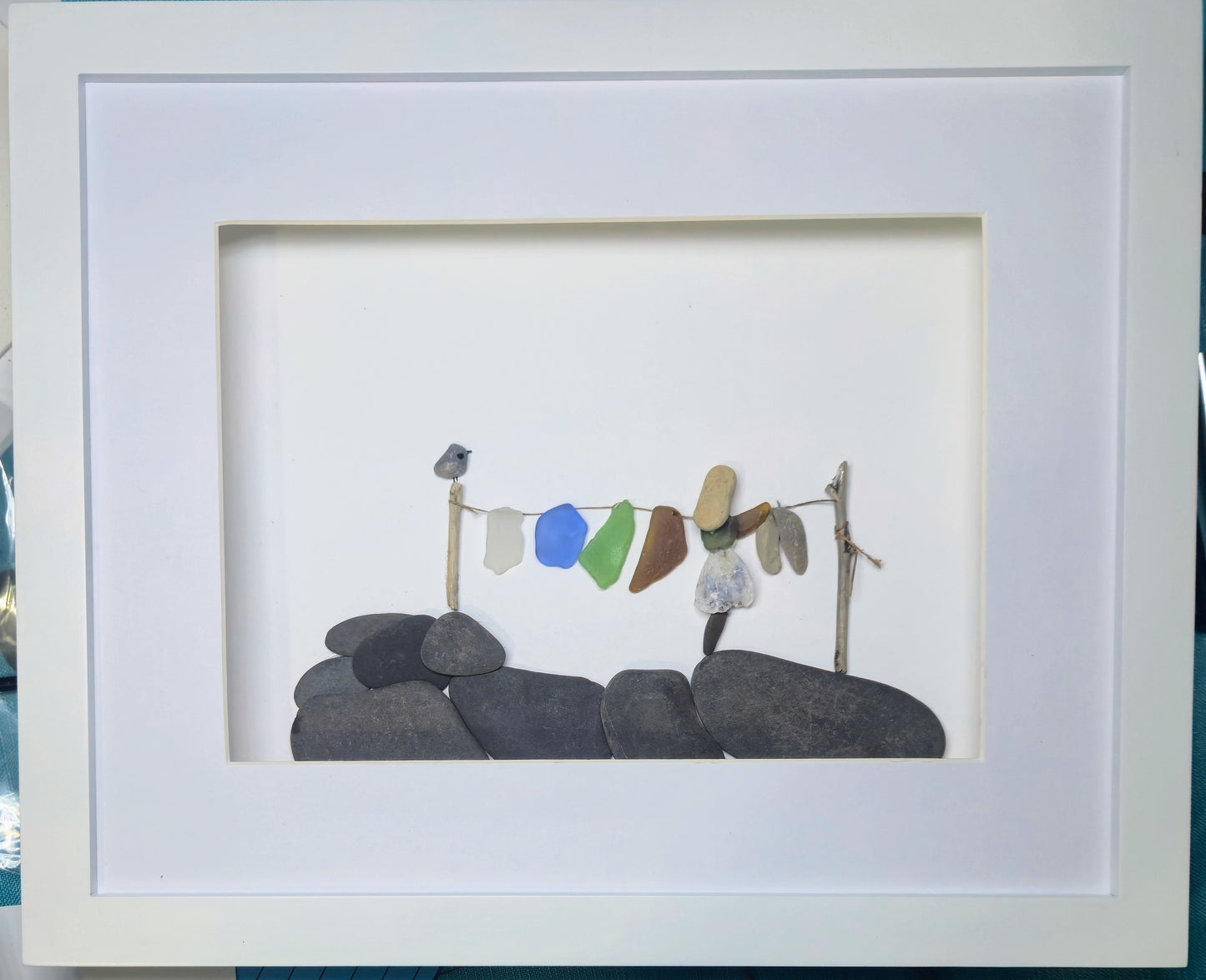 A framed sea glass and pebble art piece. Laundry day. A piece featuring small colorful stones hanging on a twig line above dark gray stones, presented in a white square frame.