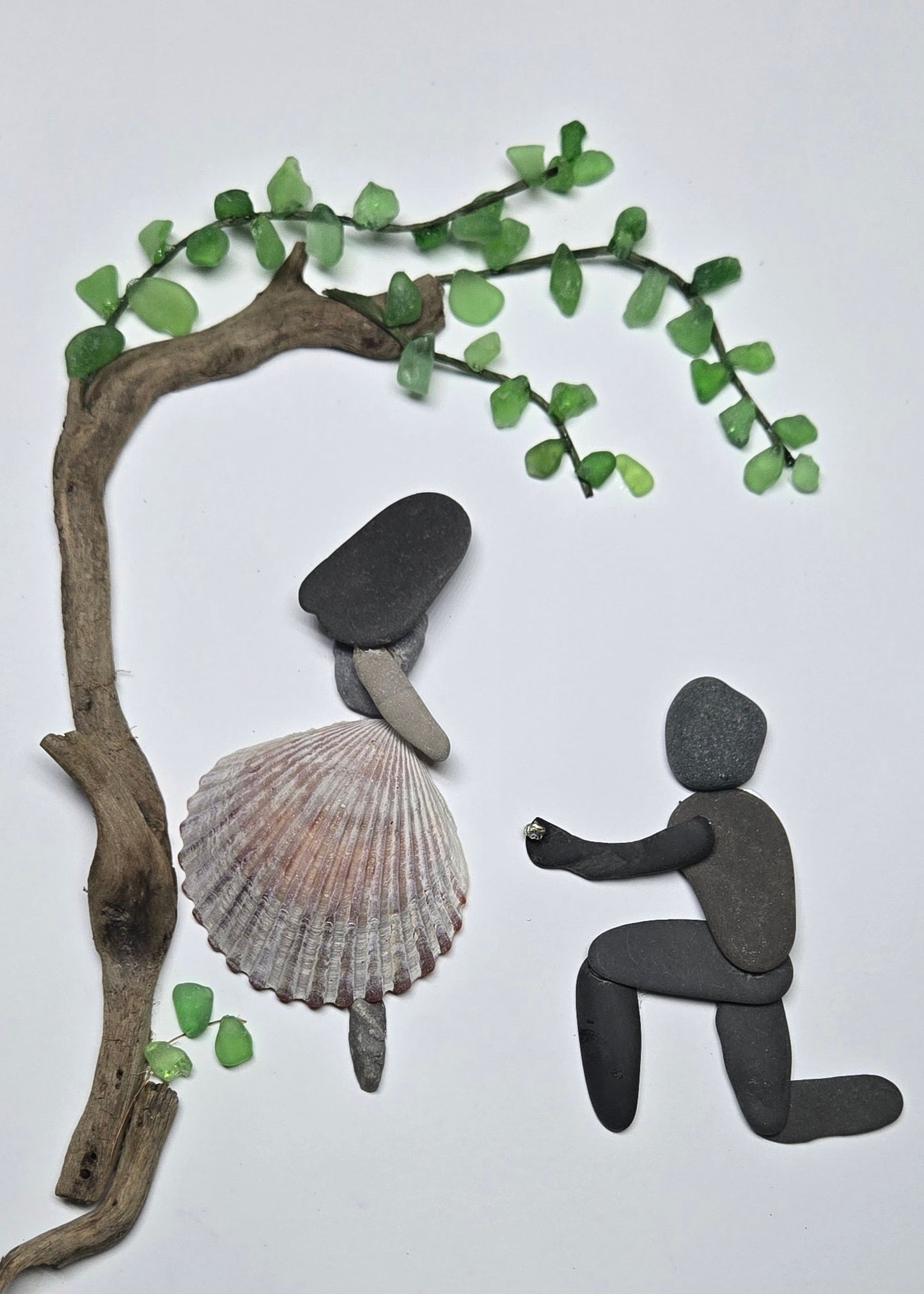 One-of-a-kind pebble art engagement scene showing a man proposing with a Swarovski crystal ring beneath a sea glass tree, with a woman wearing a shell dress from Mexico.