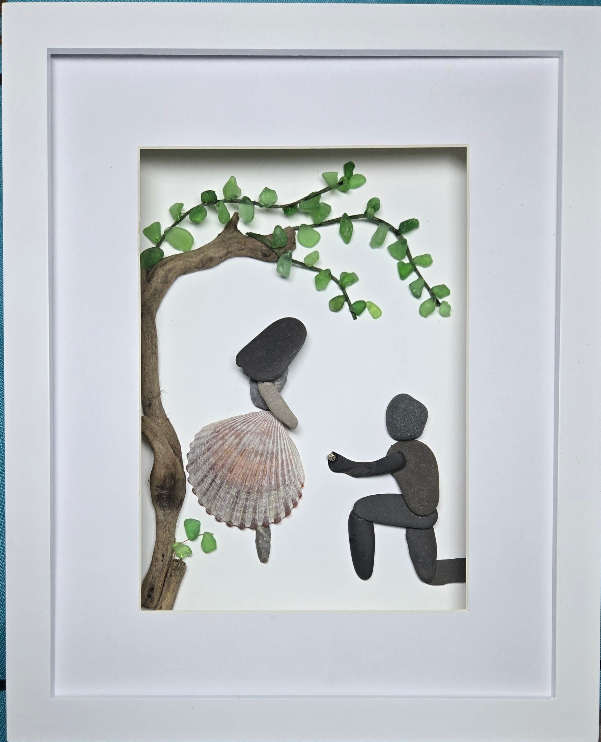 One-of-a-kind pebble art engagement scene showing a man proposing with a Swarovski crystal ring beneath a sea glass tree, with a woman wearing a shell dress from Mexico.