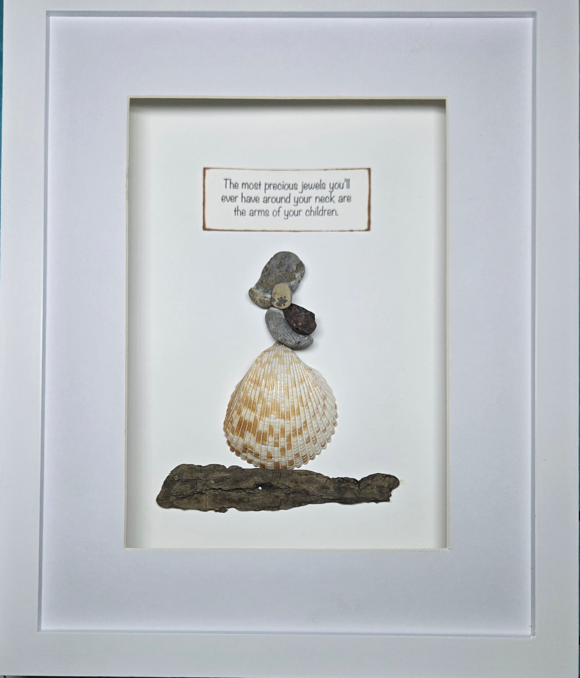 Framed wall art featuring a small balanced shell and stone arrangement on driftwood, with a small text plaque above, set inside a white frame.