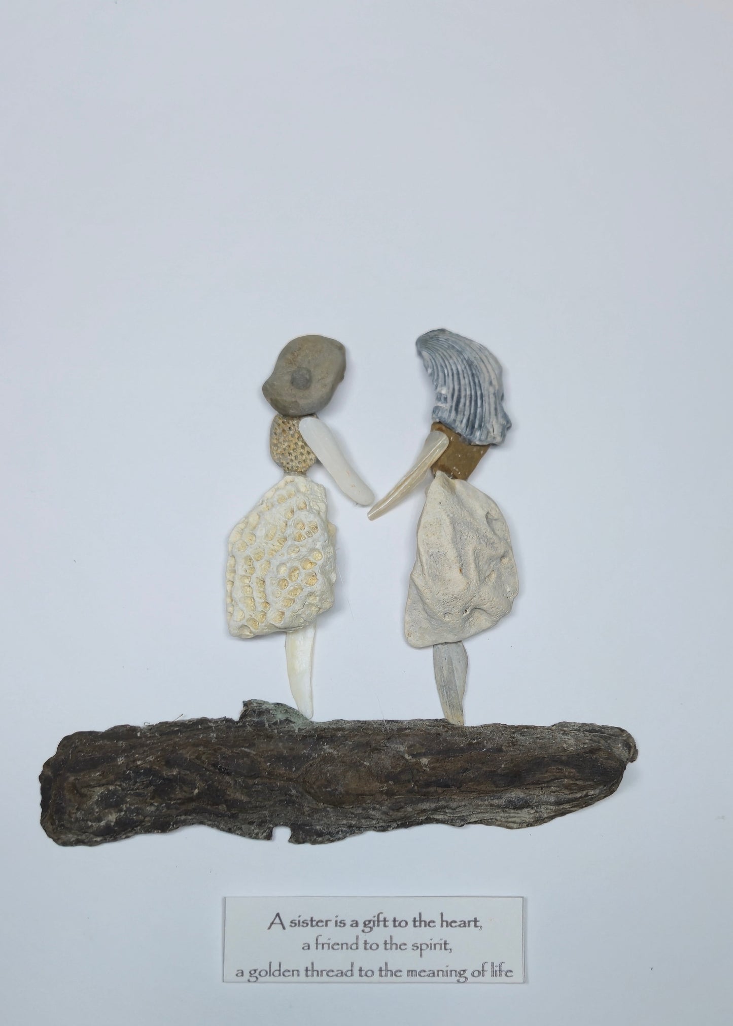 A handmade decorative sculpture of two swomen holding hands, standing on a piece of driftwood with a small caption card below. The figures are made from natural materials, including shells, bone, and textured clay, set against a plain white background.