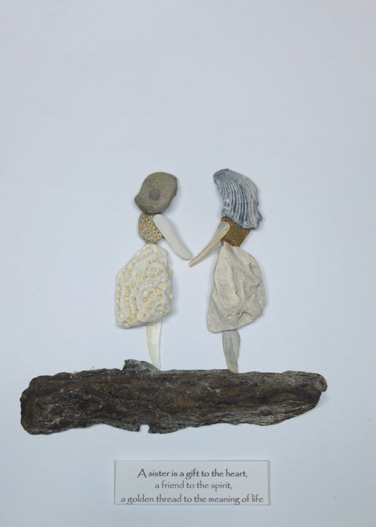 A handmade decorative sculpture of two swomen holding hands, standing on a piece of driftwood with a small caption card below. The figures are made from natural materials, including shells, bone, and textured clay, set against a plain white background.