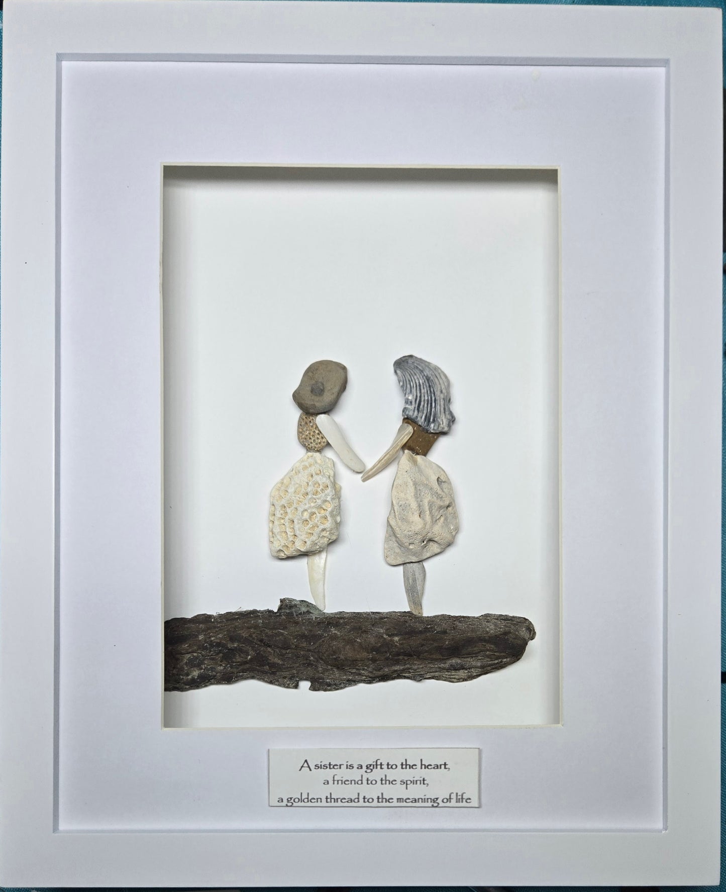 A framed handmade seashell and driftwood wall art piece featuring two ladies, a mom and a daughter or two sisters, holding hands on a log, with a white mat and a small printed quote plaque at the bottom.