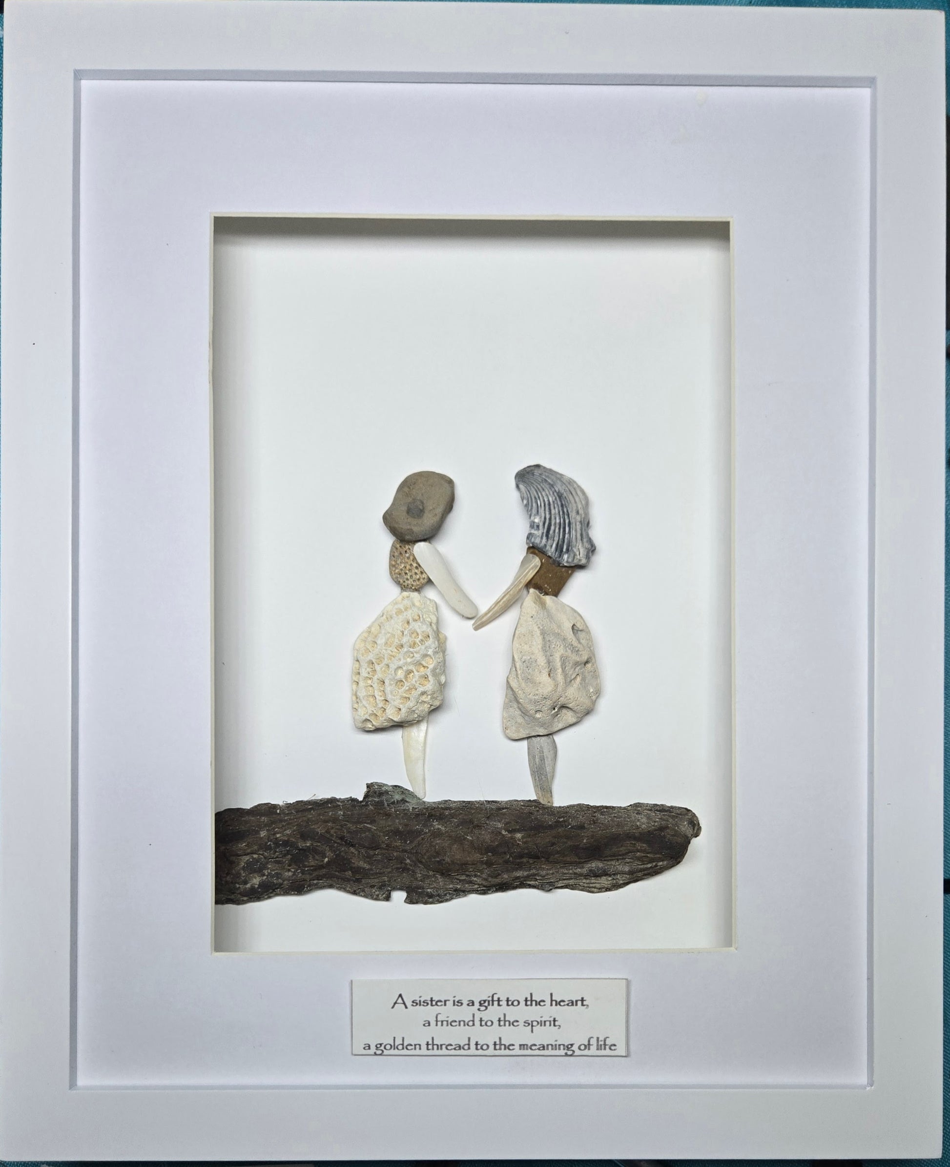 A framed handmade seashell and driftwood wall art piece featuring two ladies, a mom and a daughter or two sisters, holding hands on a log, with a white mat and a small printed quote plaque at the bottom.