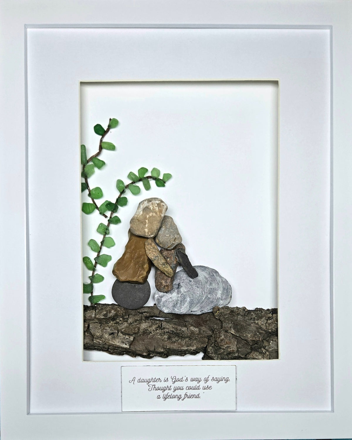 A framed decorative art piece featuring small stacked stones arranged to look like a couple embracing, with a green leafy vine on the left and a bark base. The piece is set within a wide white mat and clear outer frame, with a small printed message card at the bottom.