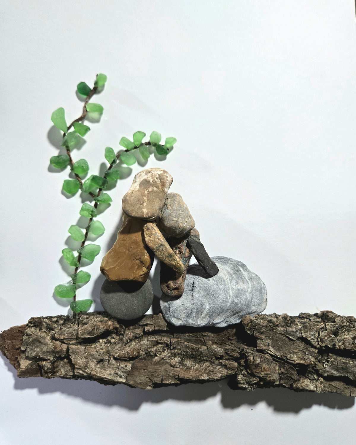 A small stack of natural stones and bark arranged on a piece of driftwood, with a green leafy twig sprig beside it, photographed against a plain white background.