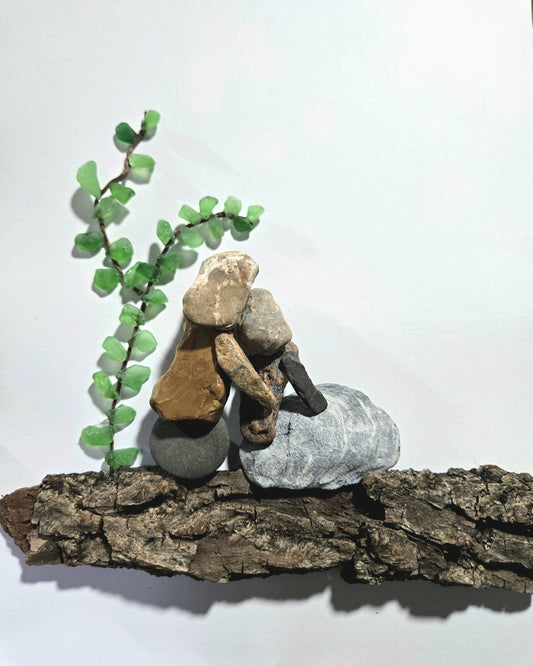 A small stack of natural stones and bark arranged on a piece of driftwood, with a green leafy twig sprig beside it, photographed against a plain white background.