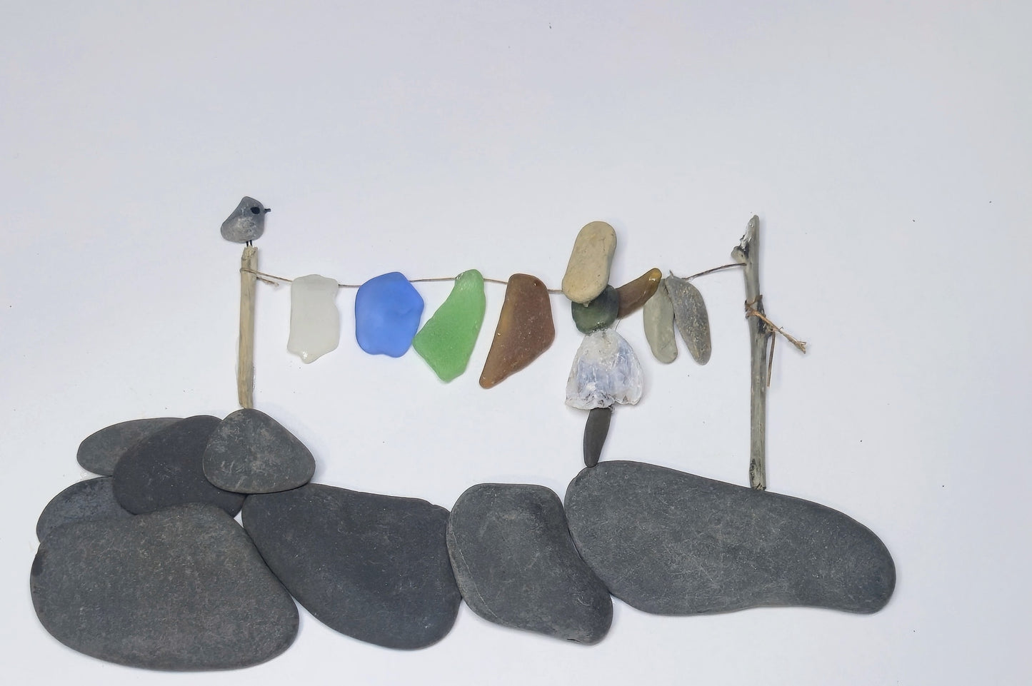 A handmade sea glass art piece depicting a small bird perched on a twig post above a string with various pieces of washed sea glass laundry set against a plain white background.