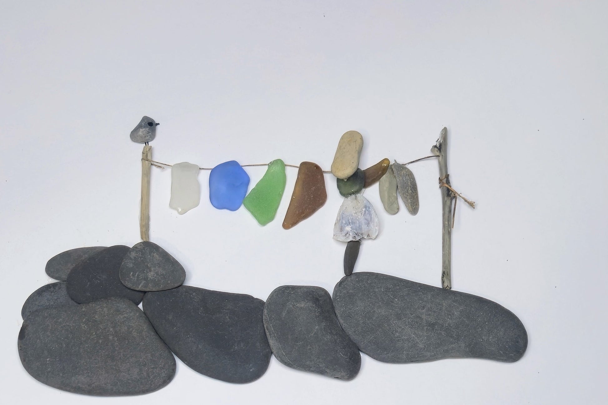 A handmade sea glass art piece depicting a small bird perched on a twig post above a string with various pieces of washed sea glass laundry set against a plain white background.