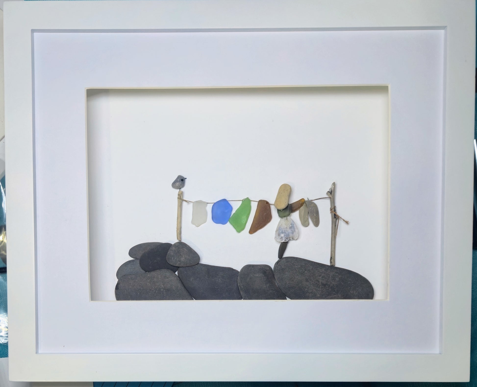 A framed sea glass and pebble art piece.  Laundry day.  A piece featuring small colorful stones hanging on a twig line above dark gray stones, presented in a white square frame.