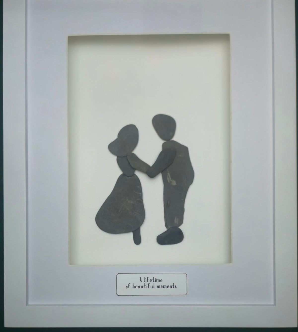 A framed minimalist art print featuring dark charcoal silhouettes made with unaltered shale stones, of a couple holding hands, set against a white rectangular print area  A small caption box at the bottom reads 'a lifetime of beautiful moments'.