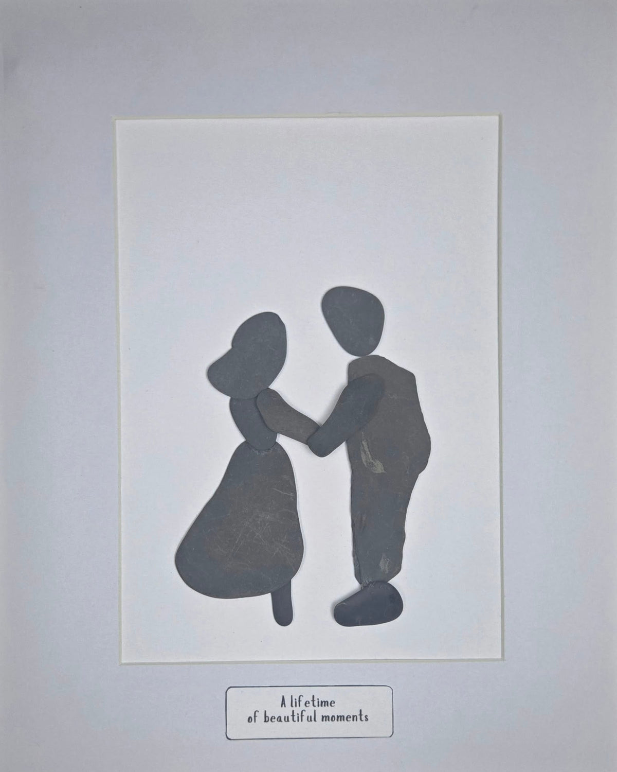 A minimalist art print featuring dark charcoal silhouettes made with unaltered shale stones, of a couple holding hands, set against a white rectangular print area  A small caption box at the bottom reads 'a lifetime of beautiful moments'.